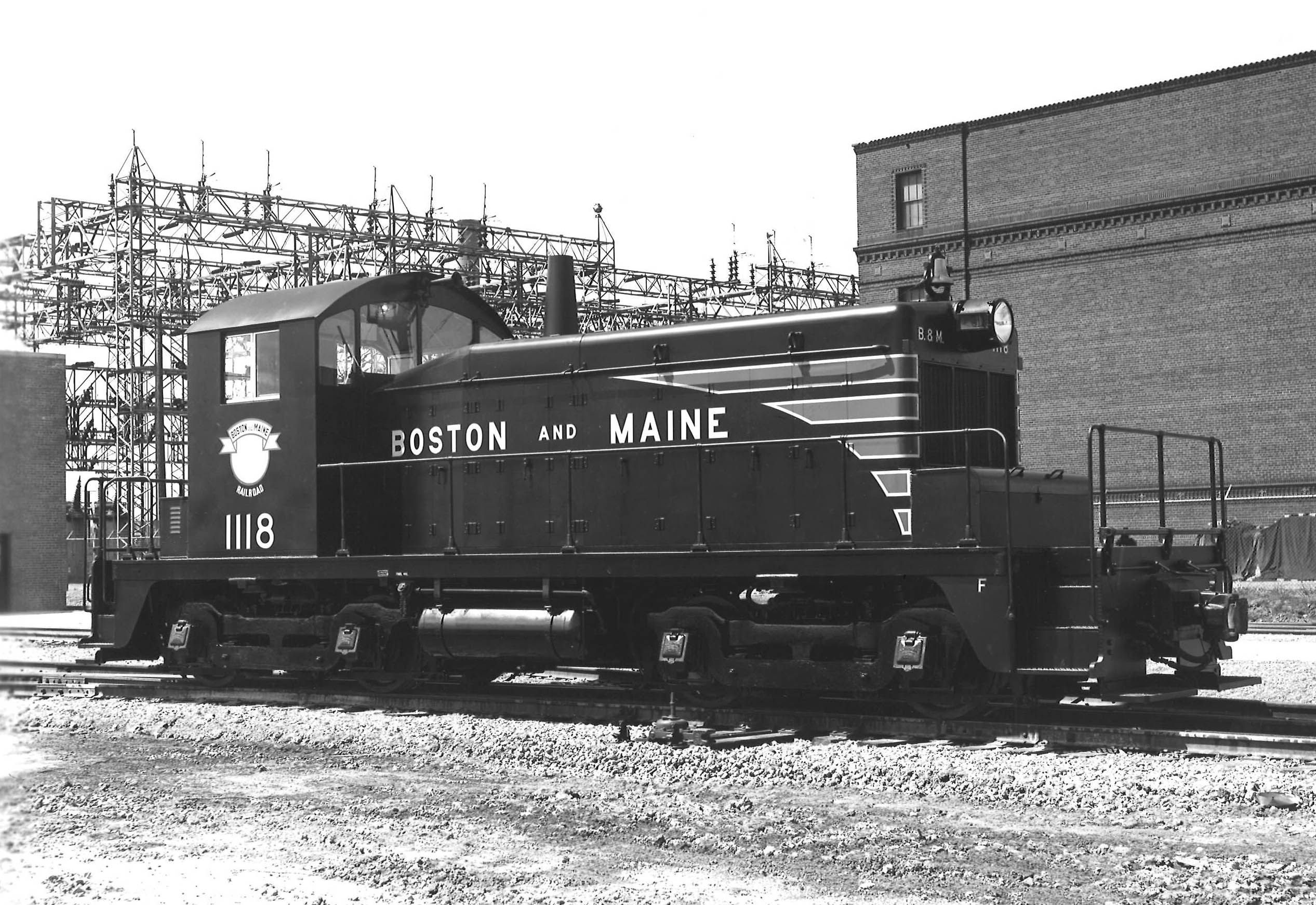 A black and white photo of an EMD switching-type locomotive sitting on the tracks in front of a building