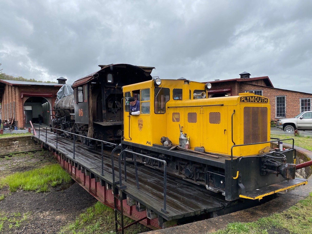 A yellow Plymouth diesel locomotive pauses on a turntable waiting to shove an East Broad Top steam locomotive into a shop.