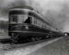 A black and white photo of a locomotive exiting from below a bridge