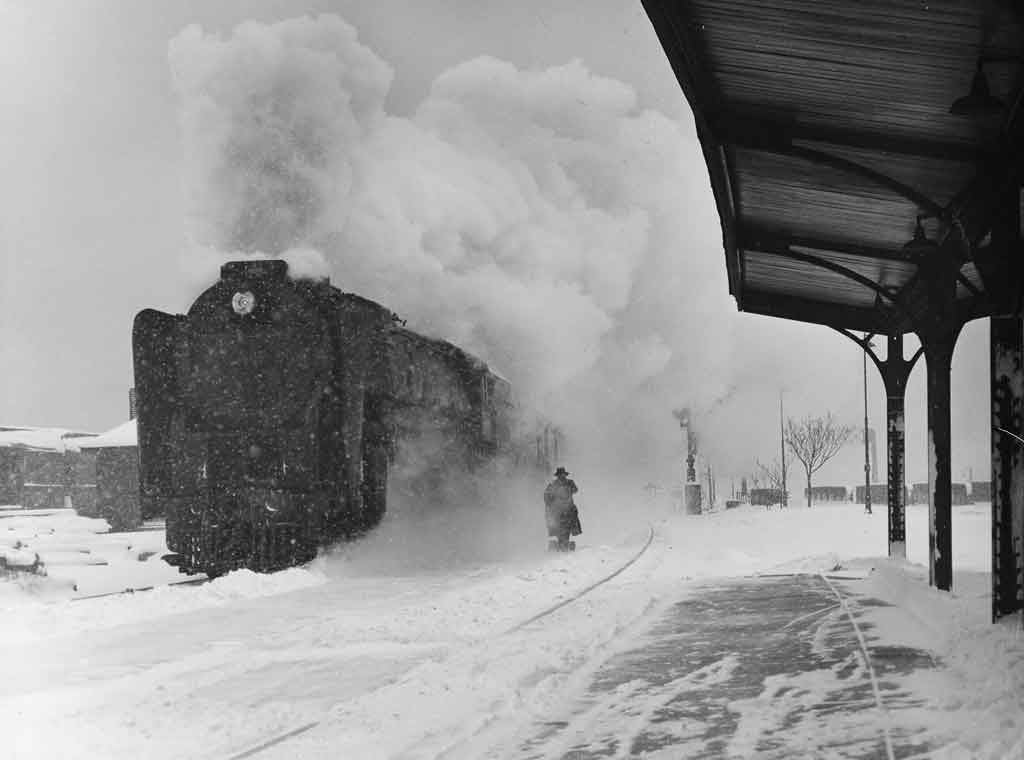 a steam engine passenger train at a passenger depot on a snowy day