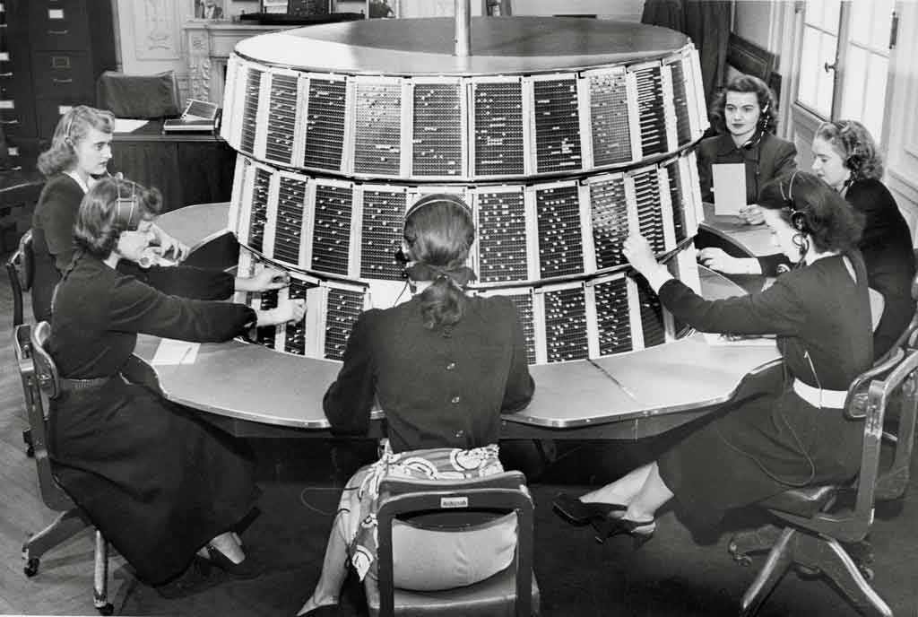 a group of women sitting at a circular table wearing headphones and plugging pegs into a switchboard