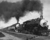 A black and white photo of two locomotives on the same track together with black smoke coming out of their chimneys 