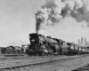 A black and white photo of a locomotive with big white smoke coming out of its chimney