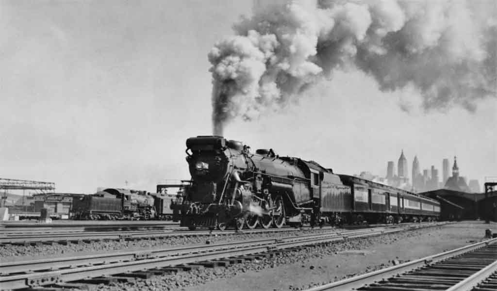 A black and white photo of a locomotive with big white smoke coming out of its chimney