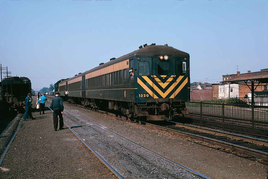 A green train with yellow stipes approaching people outside a train station