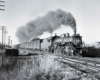 A black and white photo of a locomotive coming down the tracks with black smoke coming out of its chimney 