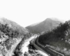 A black and white birds eye view of a train moving through mountains 