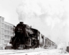 A black and white photo of a train with white smoke coming out of its chimney as it passes a building