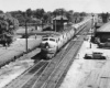 A black and white photo of a locomotive passing people and cars