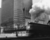 A black and white photo of a locomotive with white smoke coming out of its chimney while passing by a city