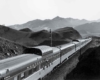 A black and white picture taken overhead a long locomotive moving through the mountains