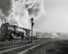 A black and white photo of a locomotive with big white smoke coming out of its chimney