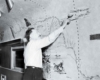 A black and white photo of an artist pointing at something on his mural inside a passenger car