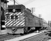 A black and white photo of a stiped locomotive passing by buildings