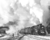 A black and white photo of a train in front of a building with big white smoke coming out of its chimney