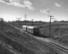 A black and white photo of a passenger train rolling down tracks near a hill