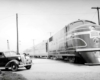 A black and white photo of a train passing by a car and young boy