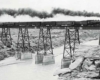 A black and white photo of a train with black smoke coming out of its chimney passing over a river