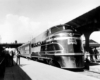 A black and white photo of a train parked at a station in front of passengers