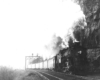 A black and white photo of a train with big white smoke coming out of its chimney as it passing by a stop light