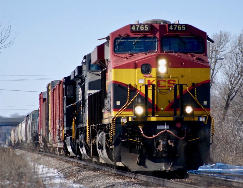 An images of a Kansas City Southern locomotive hauling a train in sunlight with blue skies in winter time.