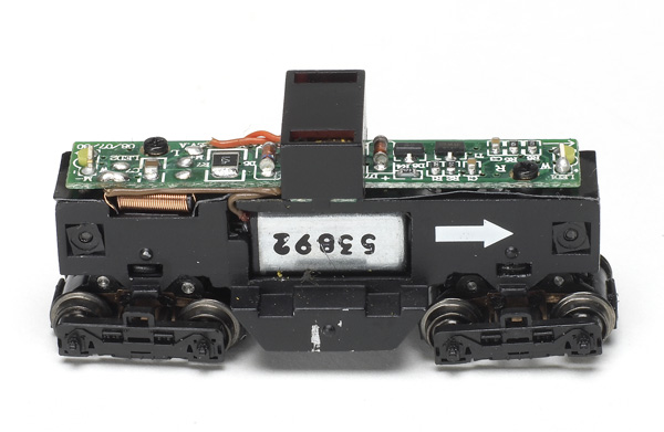 The Digital Command Control decoder is mounted on top of a split die-cast metal frame. The white arrow indicates the front of the frame.