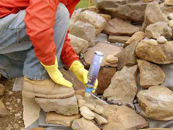 hand with rubber gloves applying sealant to rocks: How to build a pondless waterfall