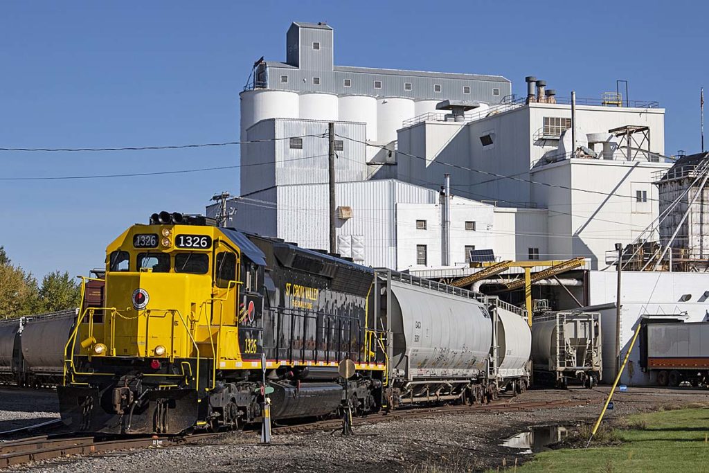 Black and yellow diesel locomotive with covered hopper cars at plant.