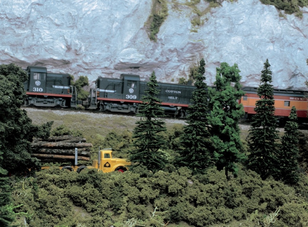 S-gauge locomotives appear to ascend uphill pulling a train to the left while a scale truck with logs moves to the right in a mountain and forest scene.