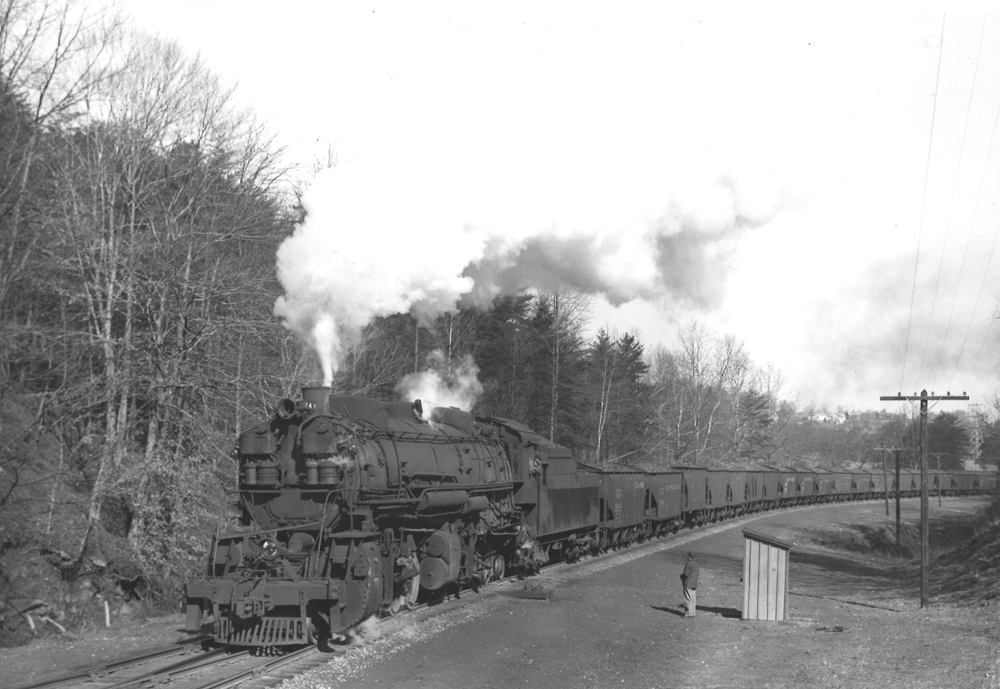 A black and white picture of someone standing on the side of the tracks watching 2-8-8-2 Mallet 740 move down the tracks with smoke coming out of its chimney