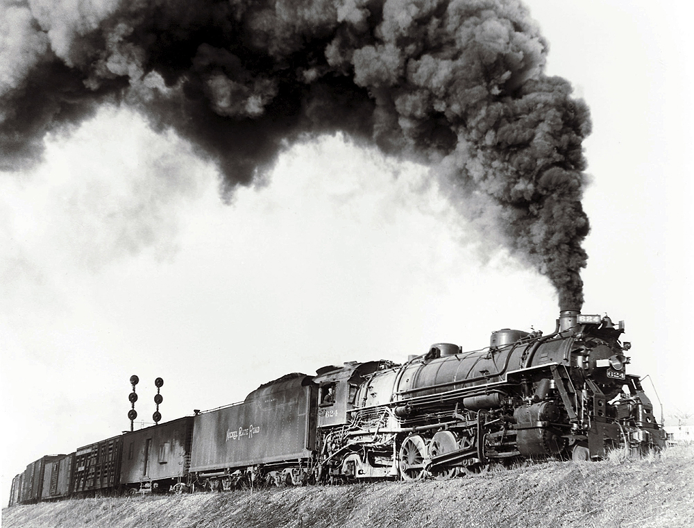 A picture of a train passing by a stoplight with big black smoke