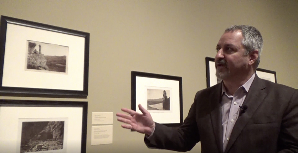 A man gestures to framed pictures on a wall.