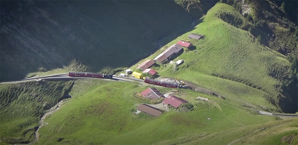 Distant steam locomotive-hauled passenger trains atop a lush green Swiss mountain.