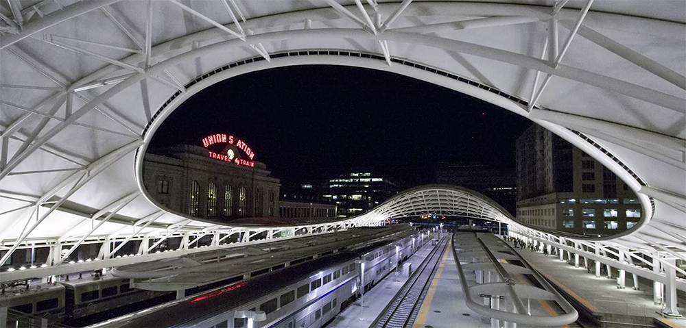 A large bean-shaped opening in a station platform roof reveals Denver Union Station's sign lit at night.