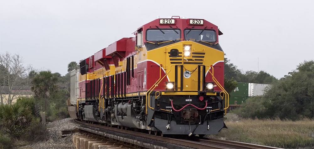 A red-and-yellow-painted Florida East Coast locomotive passes through green vegetation under cloudy skies.