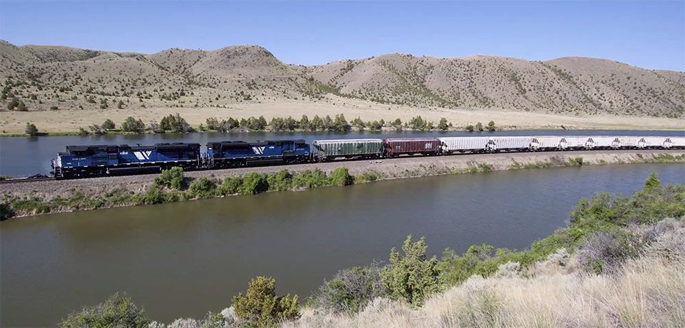 Blue-painted locomotives lead a freight train by a river under blue skies with a mountainous background.