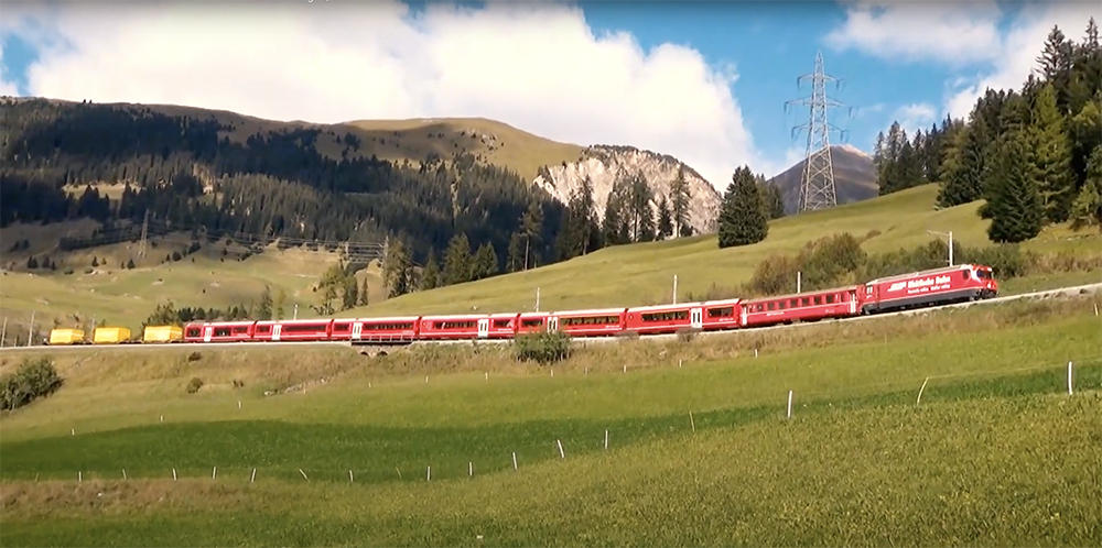 A red-painted passenger train passes through green meadows on the slopes of a mountain.