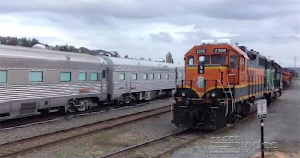 An orange-painted locomotive near silver-colored passenger cars in a rail yard.