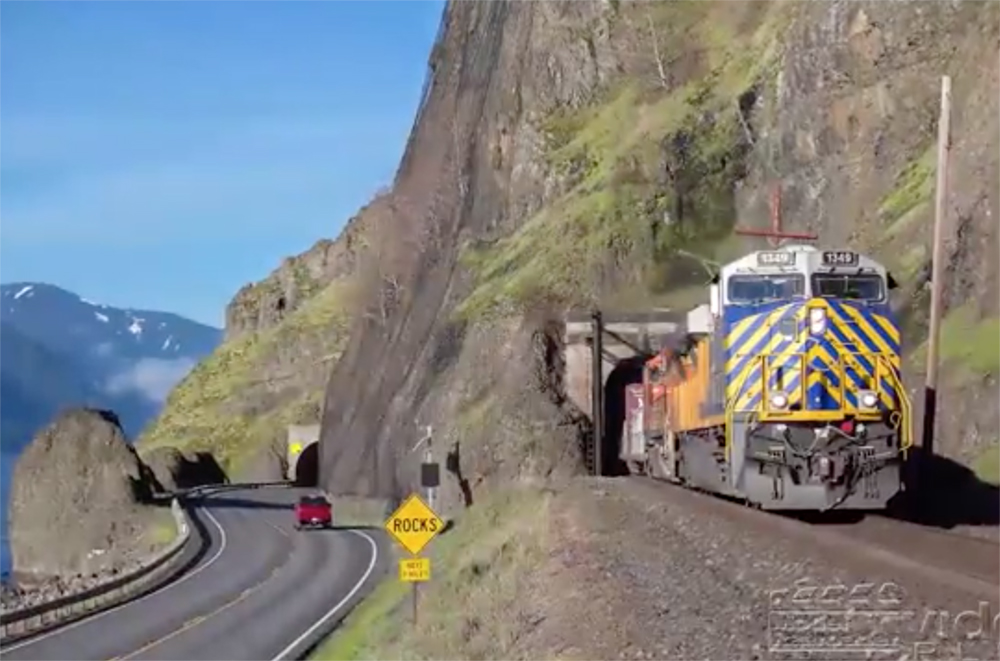 A diesel locomotive pulls a train on a track, on a ledge in mountainous terrain, above a multi-track main line.