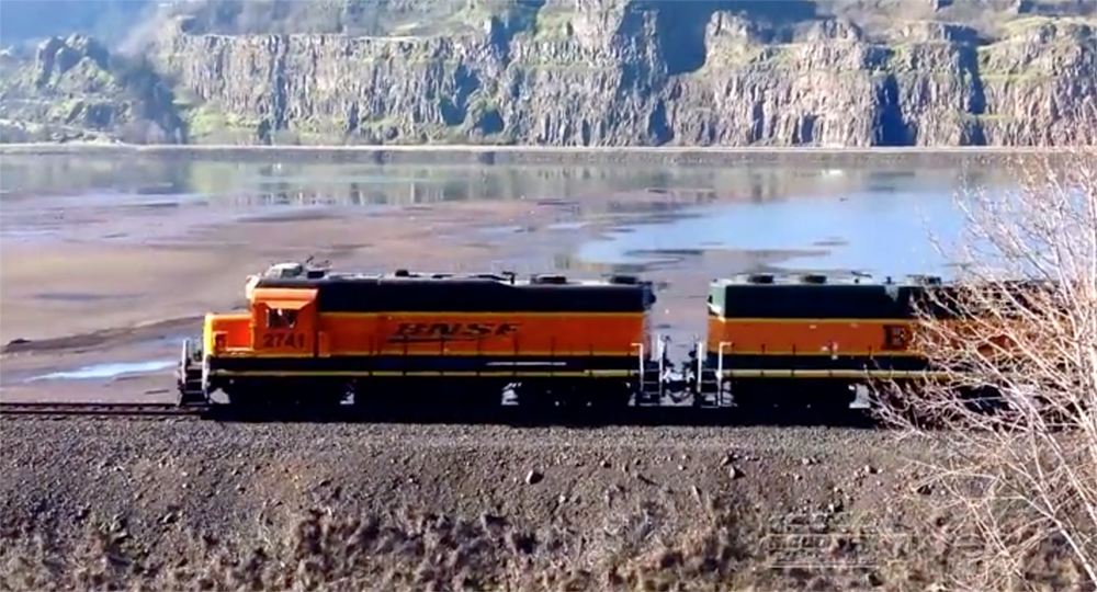Orange-painted locomotives face into low-angled sunlight with a river and rocky shoreline in the background.
