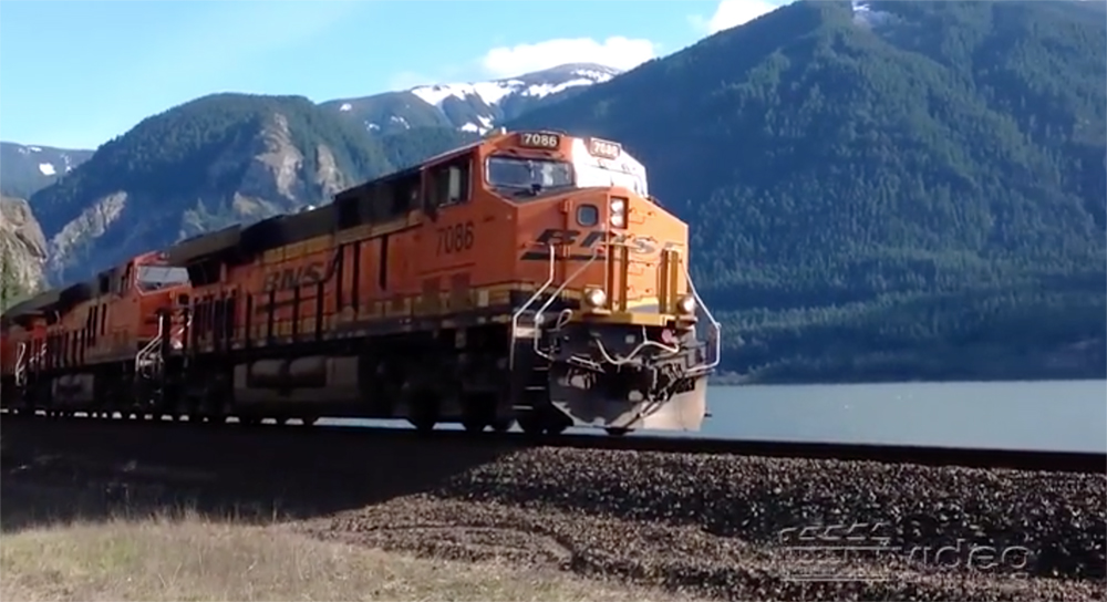 An orange-painted locomotive bathed in low angle sunlight with rugged mountains and a river in the background.