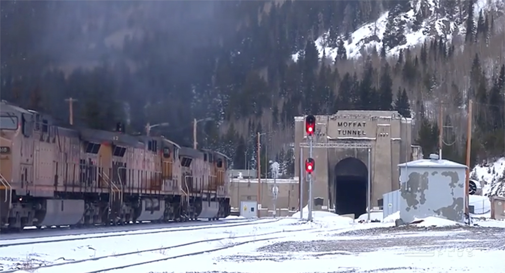 Diesel locomotives shed exhaust in misty clouds near a tunnel entrance in a snowy mountain scene.