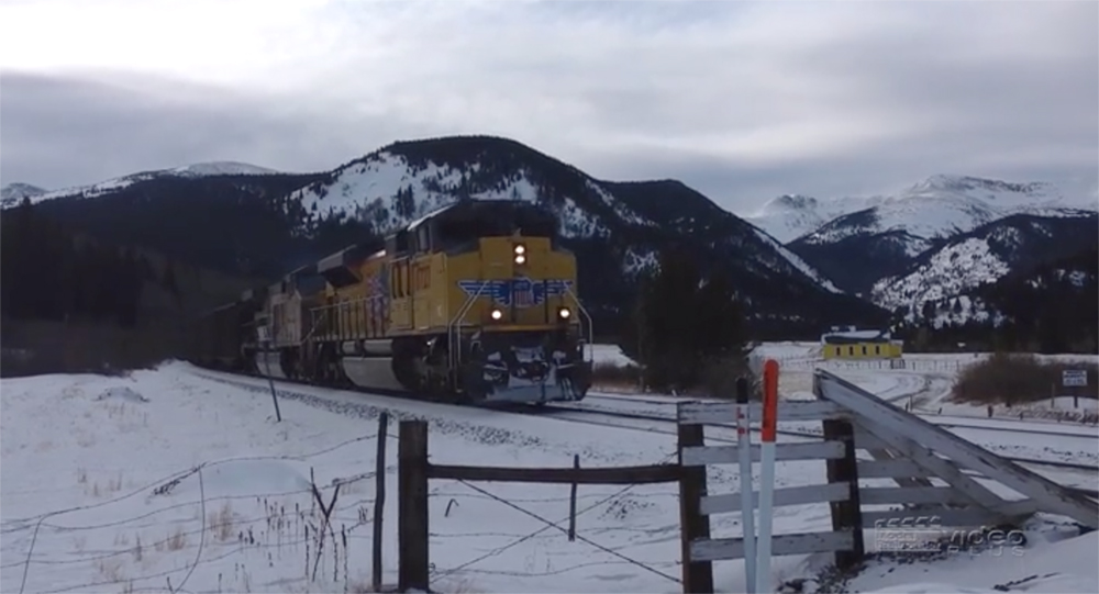 A yellow-painted locomotive travels through a snowy mountain scene with clouds and little reflected light.