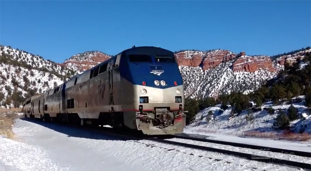 A silver-and-blue painted Amtrak diesel locomotive moves through a treeless snow covered mountain landscape under clear skies.
