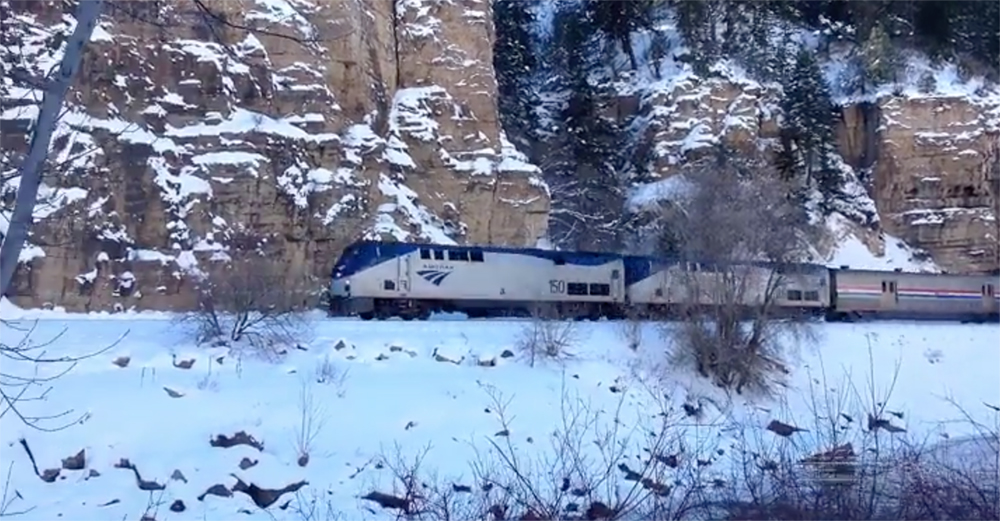 A blue-and-silver-painted Amtrak diesel locomotive leads a passenger train on a shelf with track through a snowy mountain scene.