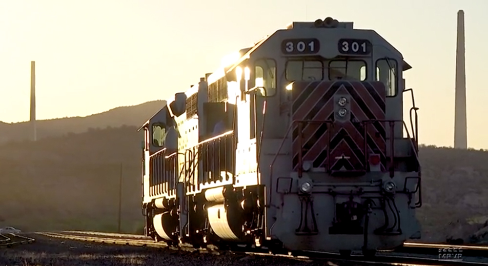 Two diesel locomotives appear backlit in a rail yard with sunlight glinting off from a low angle.