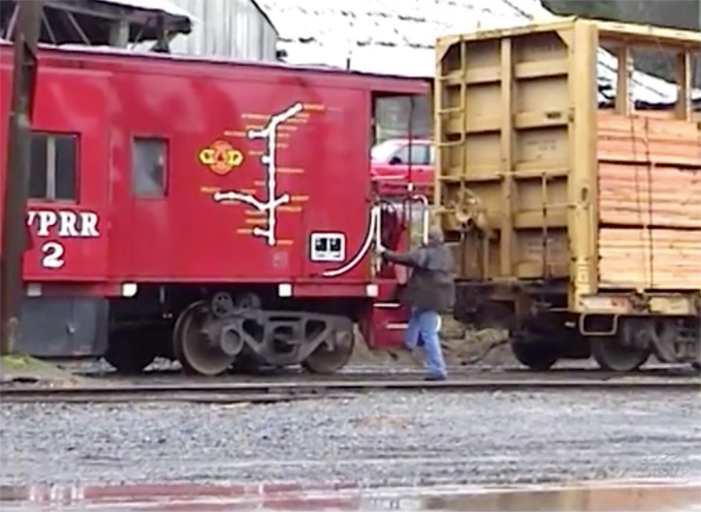 A rail worker steps onto the steps of a red-painted caboose in a rail yard.