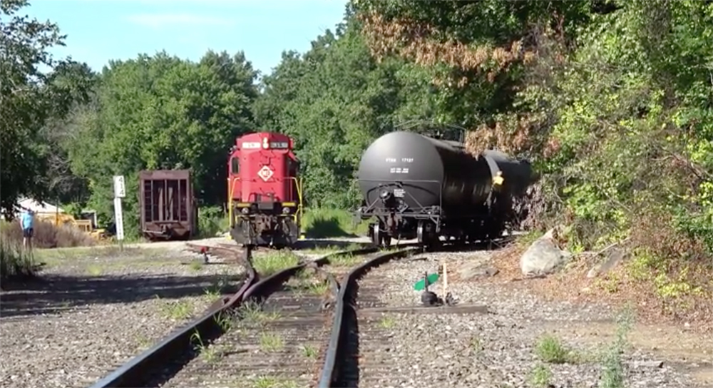 A red-painted diesel locomotive and a tank car appear in the distance on a shortline railroad's branch line.