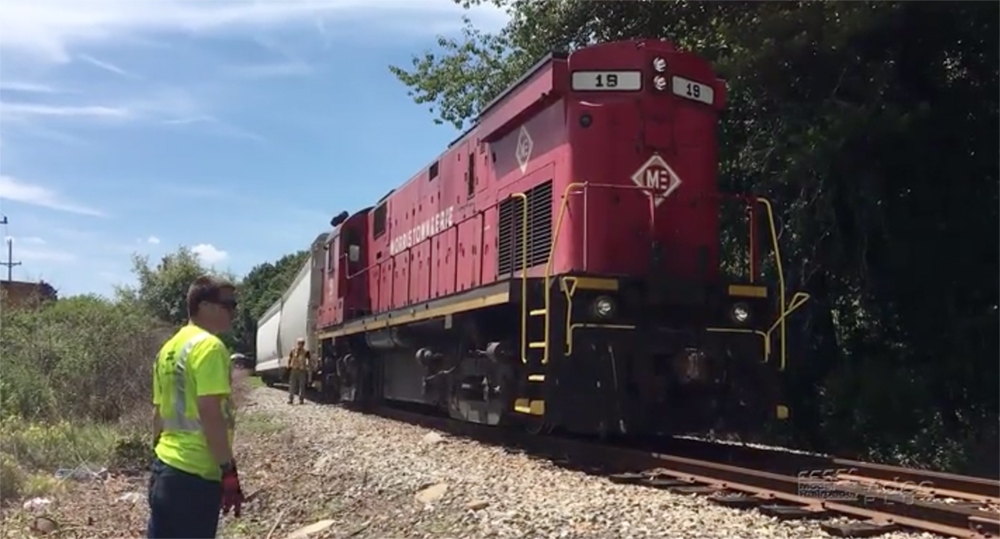 A man with high-visibility clothing stands near tracks and a red-painted diesel locomotive.
