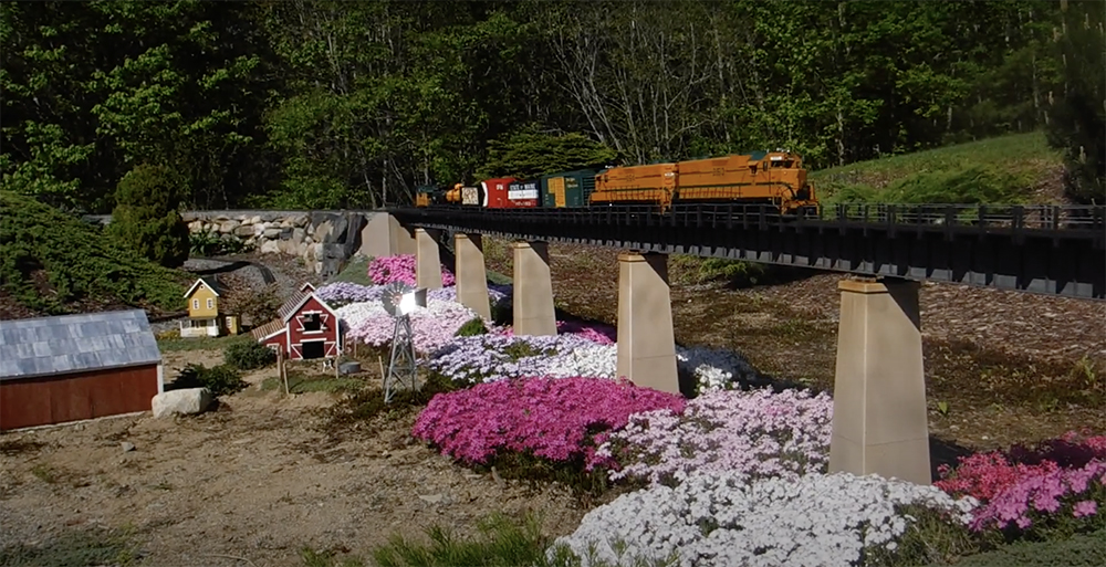 model train traveling across a bridge with flowers underneath in a garden railroad